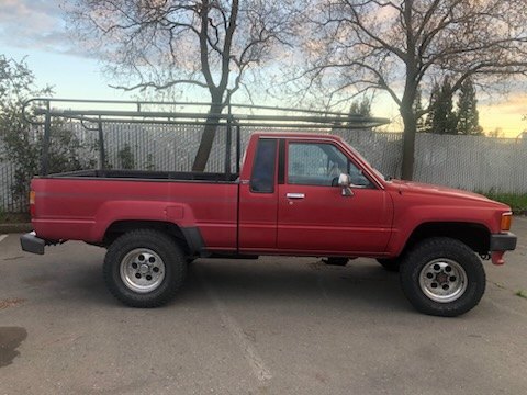 Red 1980s Toyota 4x4 pickup truck parked by a fence